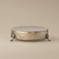 An ornate round silver pedestal stand with floral engravings and three claw feet, photographed against a solid beige background.
