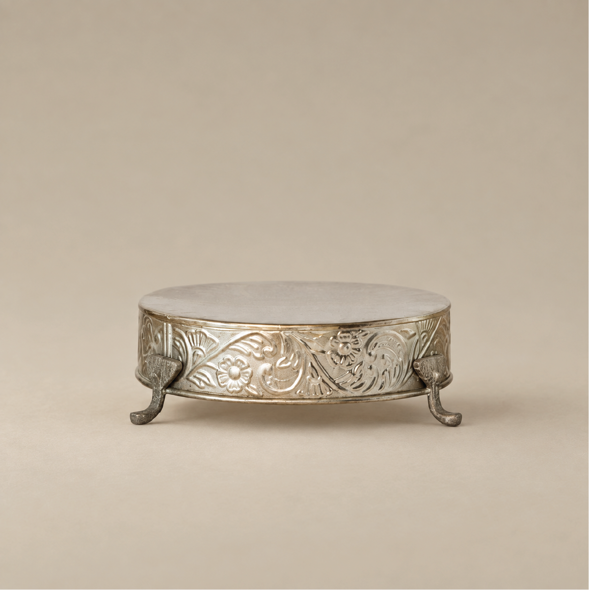 An ornate round silver pedestal stand with carved floral detailing and three small claw feet, shown on a warm beige studio background.