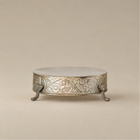 An ornate round silver pedestal stand with carved floral detailing and three small claw feet, shown on a warm beige studio background.
