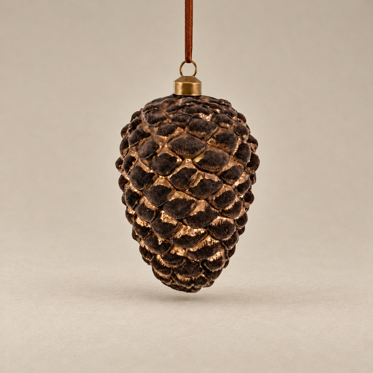 A pinecone-shaped hanging ornament with a textured dark brown and bronze finish, suspended by a brown cord with a gold cap, set against a neutral beige background.