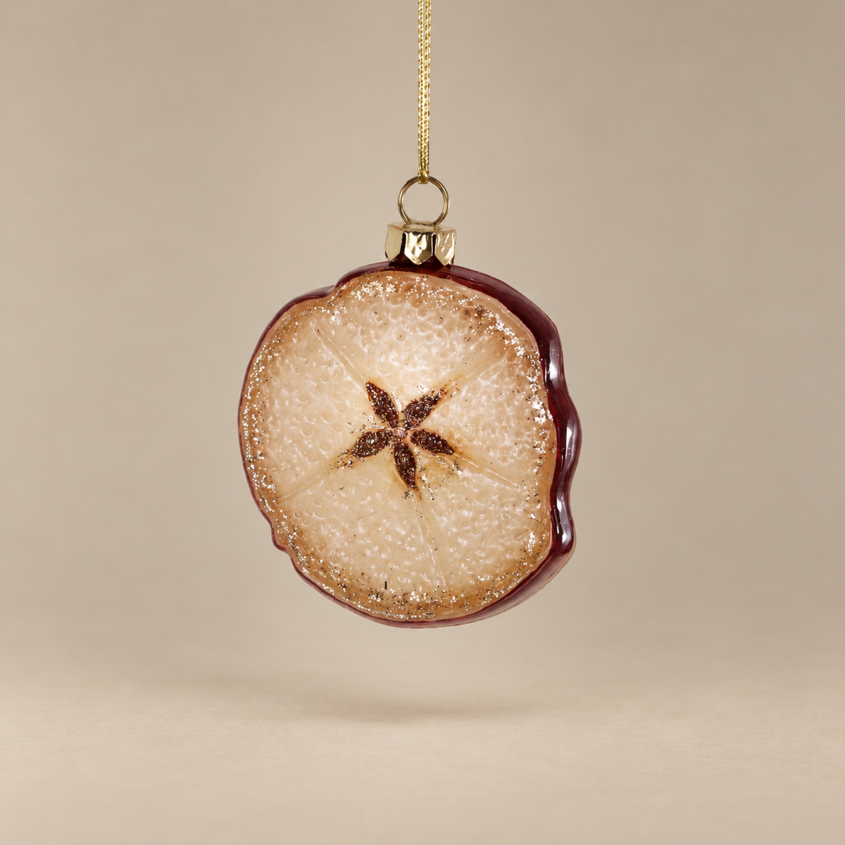 A round glass ornament shaped like a sliced apple, featuring a textured beige and brown interior with a dark brown starburst center, a dark reddish-brown outer edge, a gold hanging loop, and a gold chain against a warm beige background.