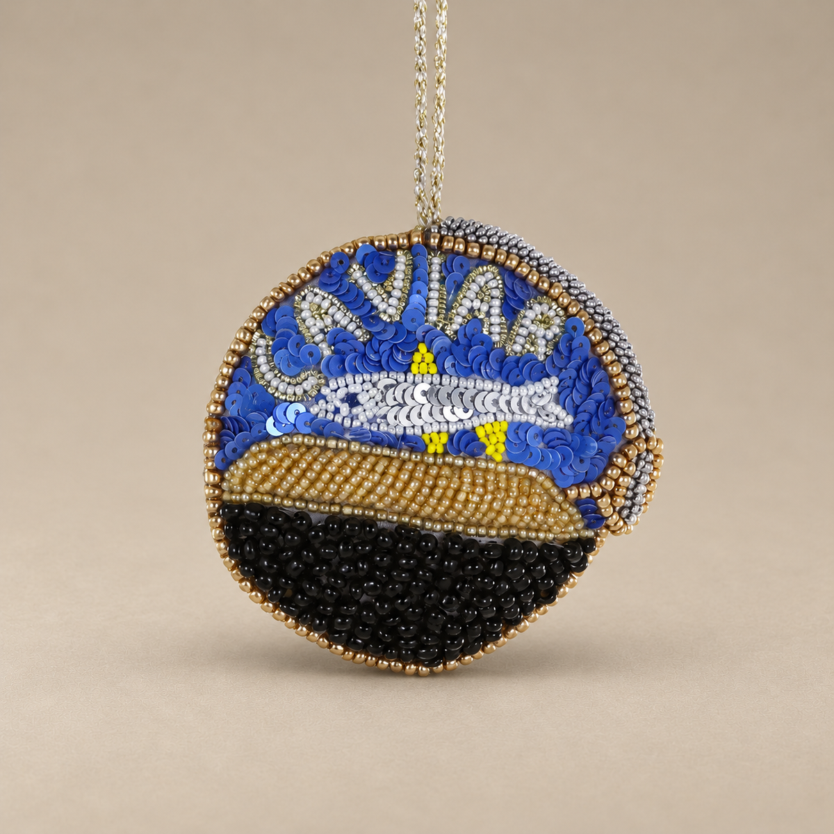 A circular beaded ornament featuring a stylized caviar design with blue, white, black, and gold beads, hanging from a silver chain against a neutral beige background.