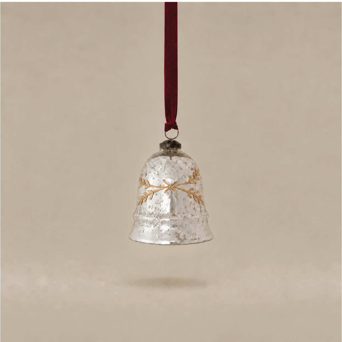 A hammered silver bell Christmas ornament with a gold wreath design, hanging from a dark red ribbon against a warm beige background.