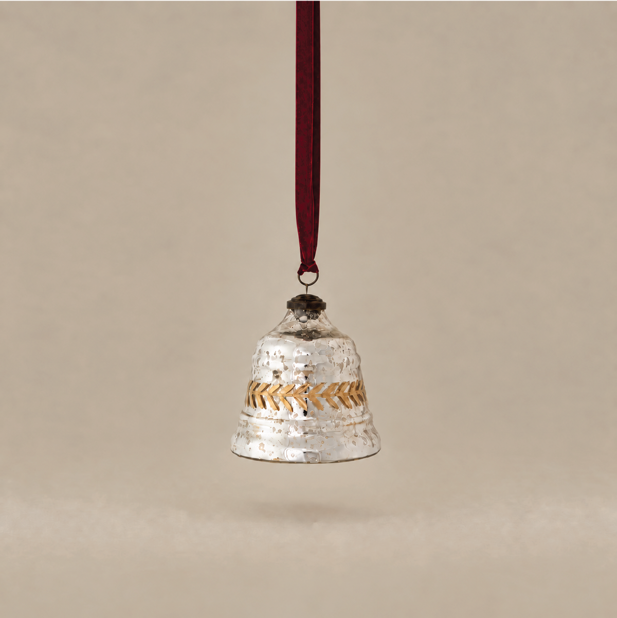 A hammered silver bell-shaped hanging ornament with a gold chevron pattern band and a dark red hanging ribbon, shown against a solid beige background.