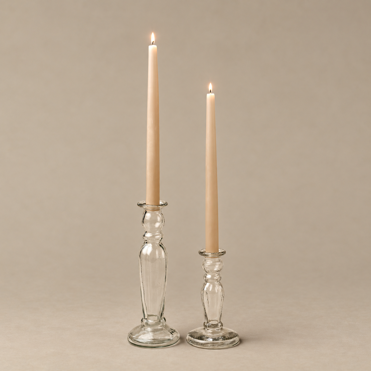Two lit taper candles in clear glass candlestick holders on a warm beige background.