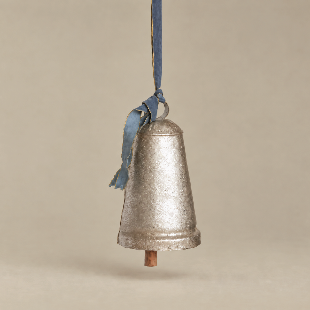 A vintage-style galvanized metal bell ornament hanging from a blue ribbon, set against a solid beige background.