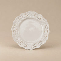 A white ceramic plate with an intricate, scalloped floral lace border, photographed against a solid beige background.