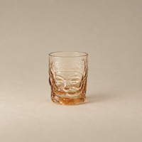 A textured clear glass tumbler with a warm amber tint, featuring a raised face design on the front, set against a solid neutral beige background.