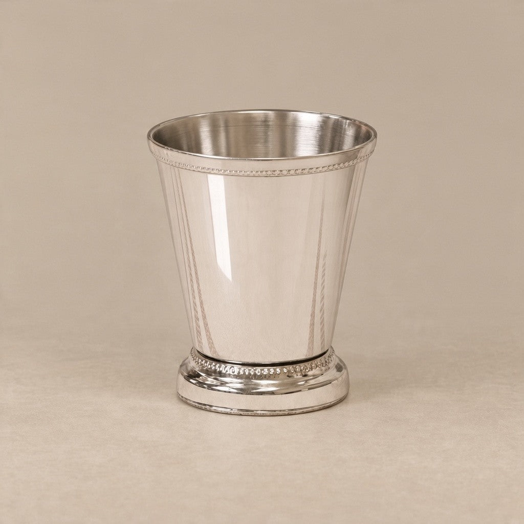 A polished silver-toned metal cup with a flared rim, vertical ribbed sides, and a decorative beaded base, photographed on a plain warm beige background.