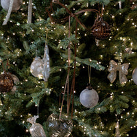 Decorated Christmas tree with warm white string lights and hanging ornaments including clear glass baubles, a silver bow ornament, a brown woven hanging ornament, and ribbons.